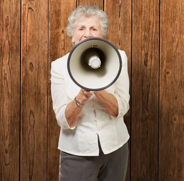 Portrait of senior woman screaming with megaphone against a wood — Stock Photo, Image