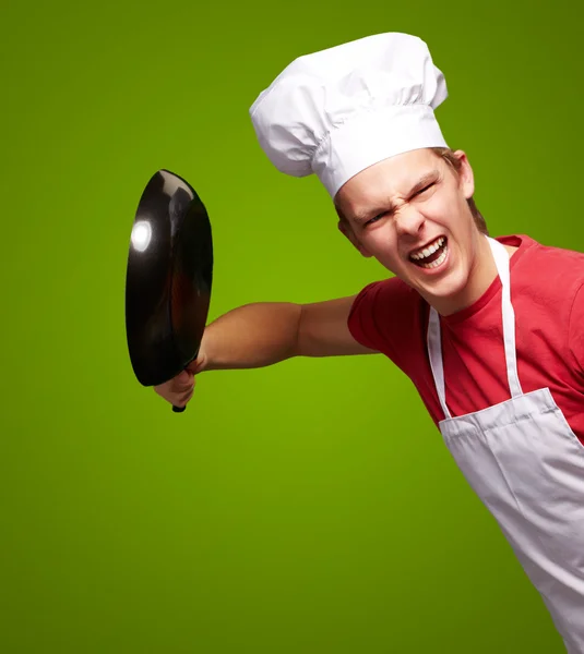 Портрет разгневанного молодого повара, бьющего сковородкой по зеленой набережной — стоковое фото
