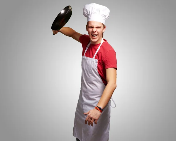Retrato del joven cocinero enojado golpeando con la sartén sobre la espalda gris — Foto de Stock