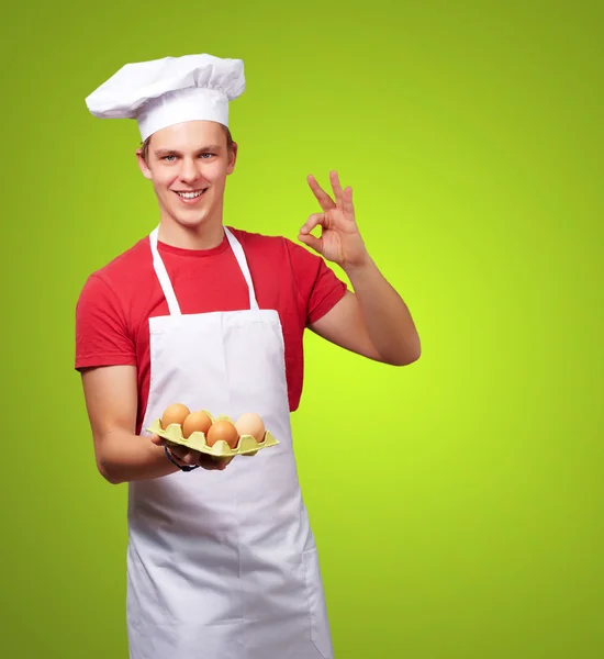 Retrato de jovem cozinheiro segurando caixa de ovo e fazendo bom gestur — Fotografia de Stock