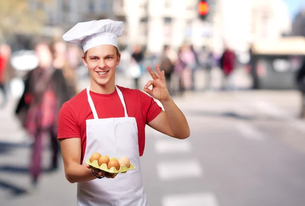 Retrato de un joven cocinero sosteniendo una caja de huevos y haciendo un buen gesto —  Fotos de Stock