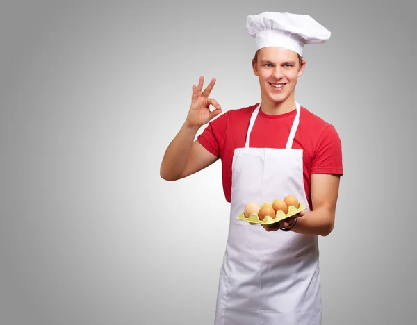 Retrato de jovem cozinheiro segurando caixa de ovo e fazendo bom gestur — Fotografia de Stock