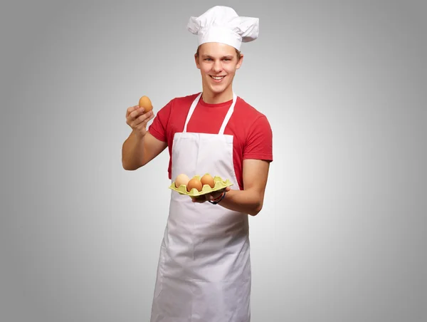 Porträtt av ung kock man hålla ägget låda över grå bakgrund — Stockfoto