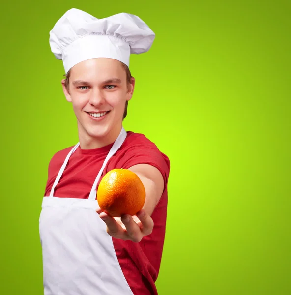 Portrait of young cook man offering orange over green background — Stock Photo, Image