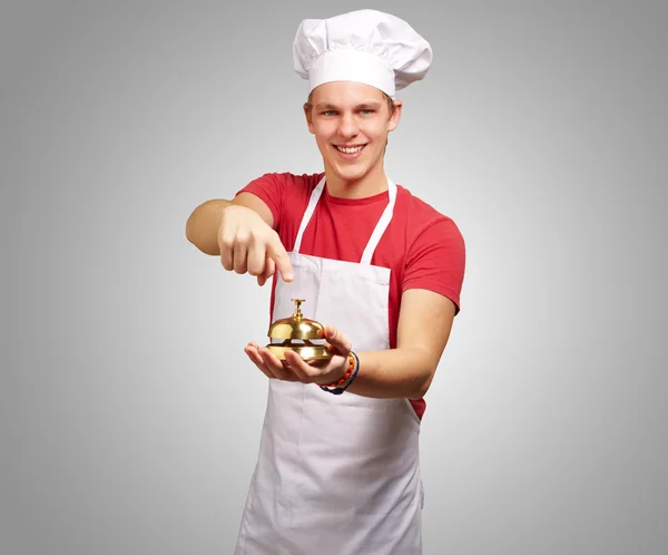Retrato del joven cocinero presionando una campana dorada sobre la espalda gris — Foto de Stock