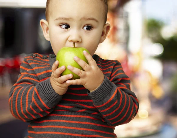 Portrait of a handsome kid bitting a green apple against a carou — Stock Photo, Image