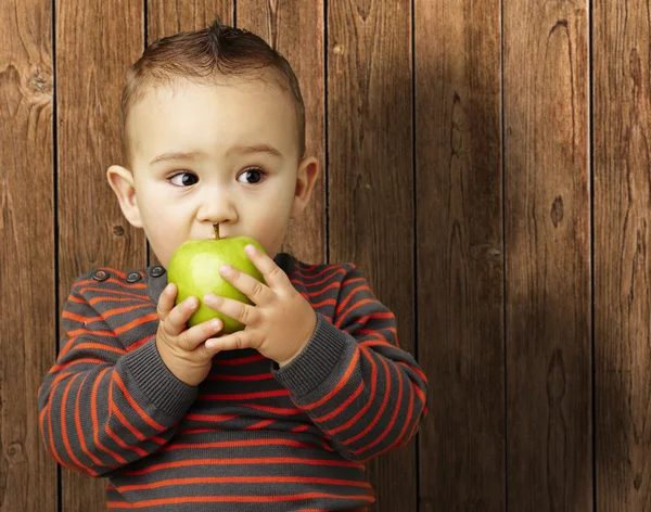 Portrait of a handsome kid bitting a green apple against a woode — Stock Photo, Image