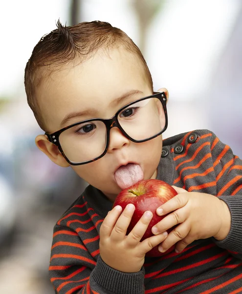 Retrato de um garoto bonito usando óculos chupando uma maçã vermelha i — Fotografia de Stock