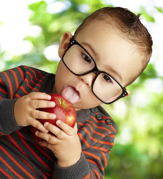 Portrait of a handsome kid wearing glasses sucking a red apple a — Stock Photo, Image