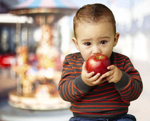 Portrait of a handsome kid sucking a red apple against a carouse — Stock Photo, Image