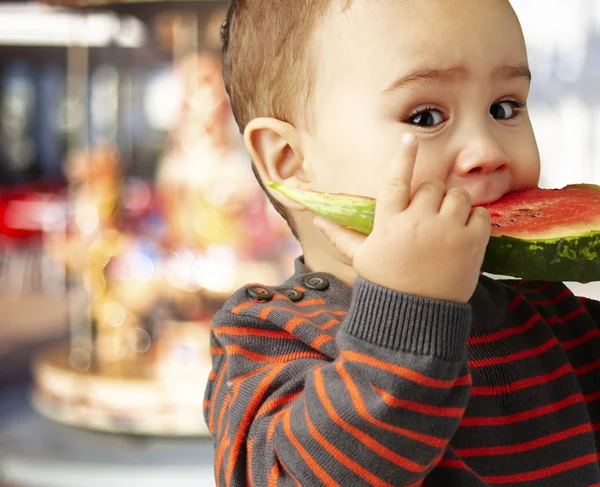 Portrét pohledný kluk bitting meloun proti carous — Stock fotografie