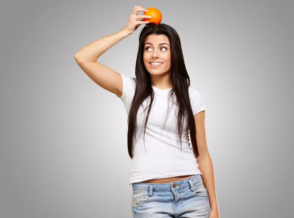 Πορτρέτο της νεαρής γυναίκας κρατώντας πορτοκαλί στο κεφάλι της, πέρα από το γκρι — Φωτογραφία Αρχείου