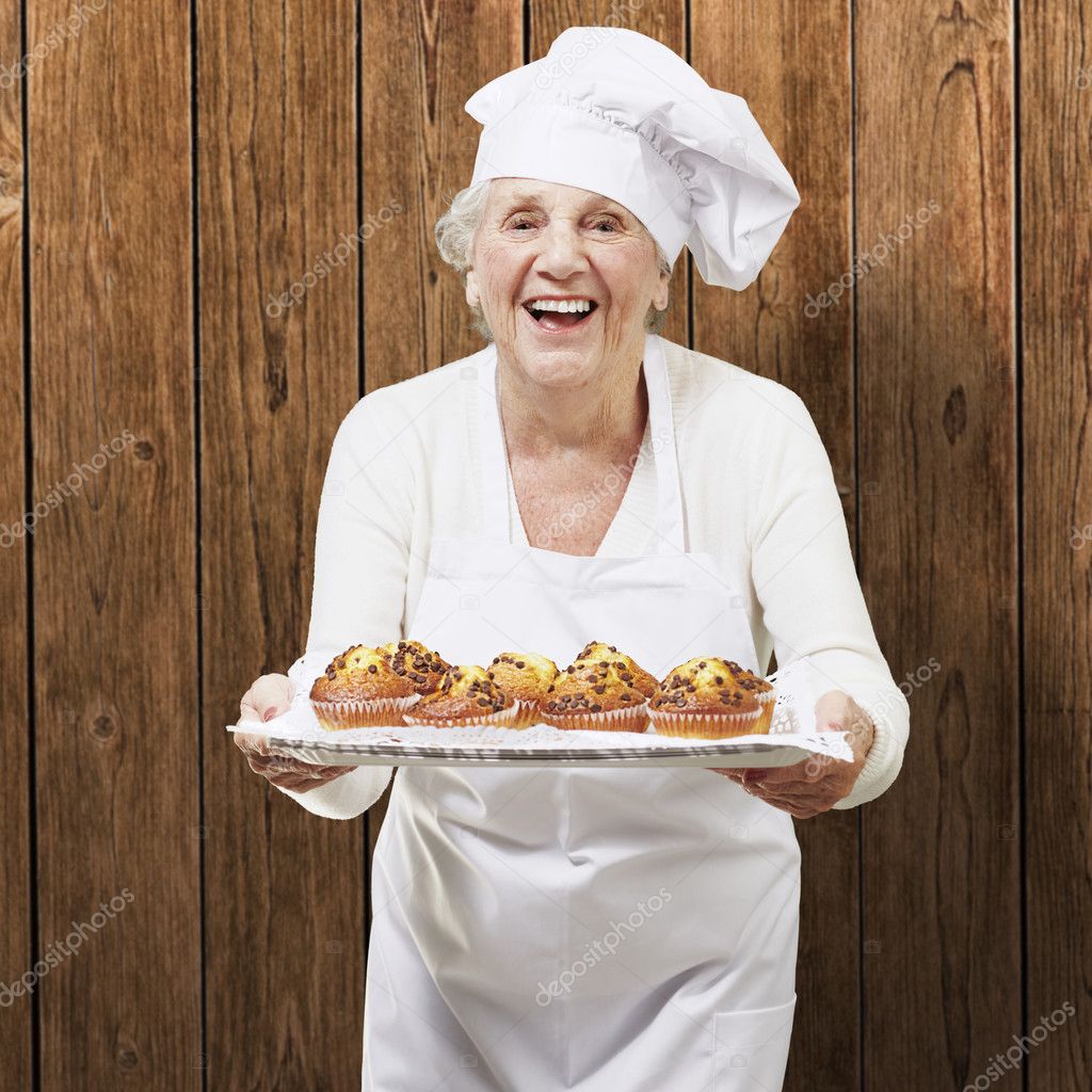 Senior woman cook holding a tray with muffins against a wooden b