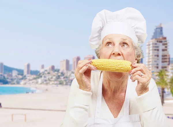 Retrato de cocinera senior comiendo mazorca de maíz contra una playa — Foto de Stock