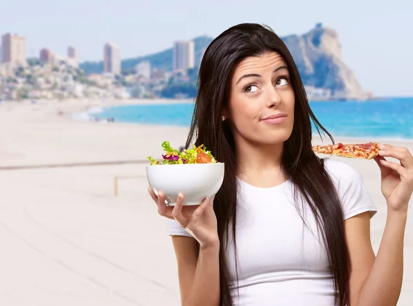 Portret van een jonge vrouw kiezen pizza of salade tegen een strand — Stockfoto