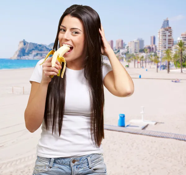 Portrét mladé ženy jíst banán proti pláži — Stock fotografie