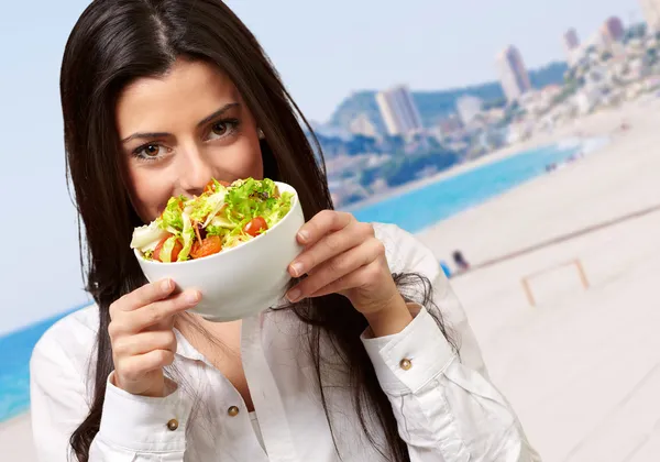 Portrait of young woman holding a fresh salad against a beach — Stock Photo, Image