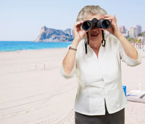Portrait of senior woman looking through a binoculars in the bea — Stock Photo, Image