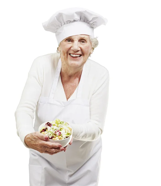 Femme âgée cuisinier tenant un bol avec salade contre un dos blanc — Photo