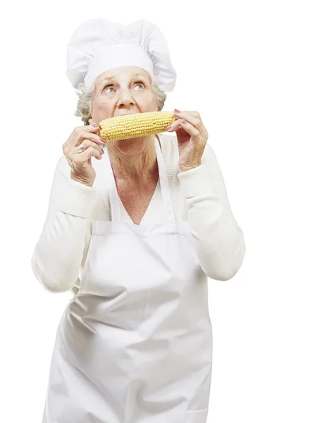 Donna anziana cuoco mangiare una pannocchia di mais su uno sfondo bianco — Foto Stock