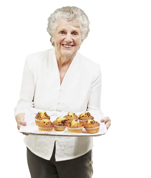Senior vrouw glimlachend en houden een dienblad met muffins tegen een w — Stockfoto
