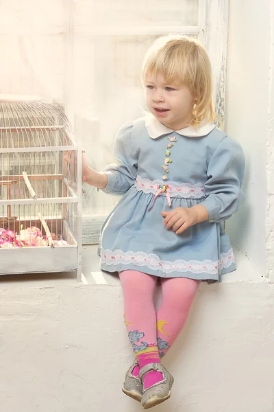 Маленька дівчинка в сукні, що сидить біля вікна з клітиною — стокове фото