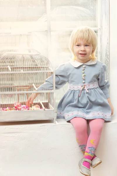 Маленька дівчинка в сукні, що сидить біля вікна з клітиною — стокове фото
