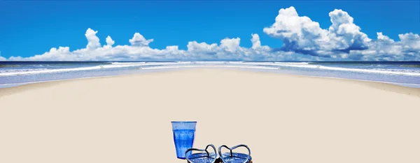 Playa tropical con zapatos de playa y vaso de agua azul —  Fotos de Stock