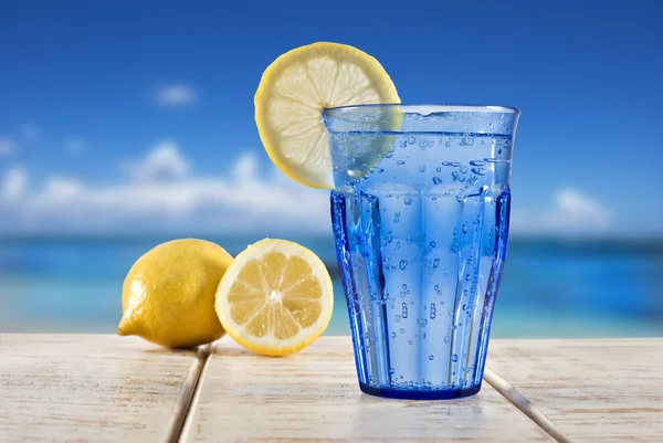 Um copo azul com água espumante e limão em um deck de madeira com vista para uma praia tropical foco no limão — Fotografia de Stock