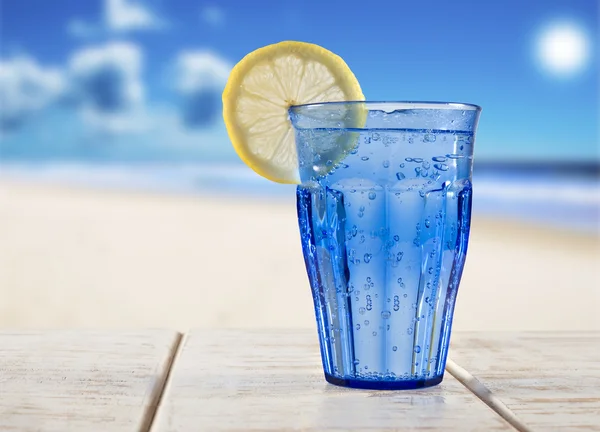 Een blauw glas met bruiswater en citroen op een houten terras met uitzicht op een tropisch strand - focus op de citroen — Stockfoto