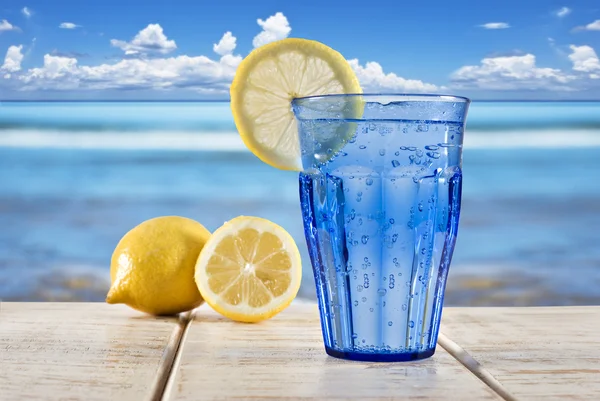 Een blauw glas met bruiswater en citroen op een houten terras met uitzicht op een tropisch strand — Stockfoto