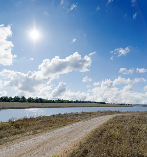 Sun on cloudy sky over rural road and river near it — Stock Photo, Image