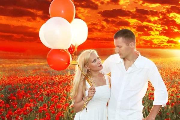 Glückliches Paar, das sich über Mohnfeld und Sonnenuntergang umarmt und Ballons in der Hand hält — Stockfoto