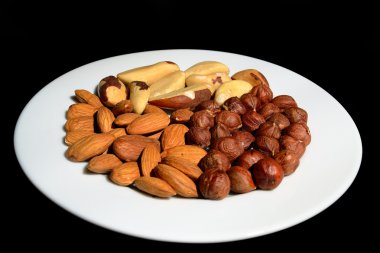 Hazelnuts, Brazil nuts, almonds, lie on the white plate on a black backgrou clipart