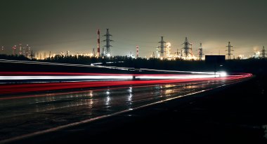 Car lights on a highway at night on the background of the industrial landscape. clipart