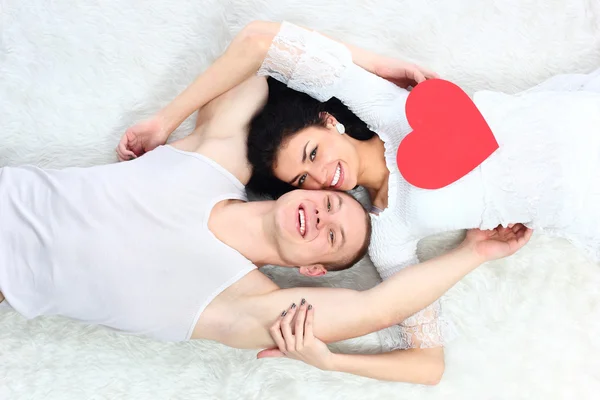 Young lovely couple holding red heart together lie in a bed — Stock Photo, Image