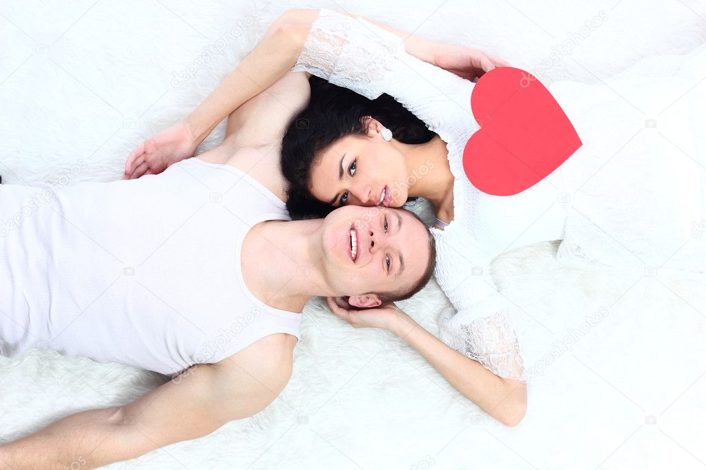 Young lovely couple holding red heart together lying in a bed, valentine