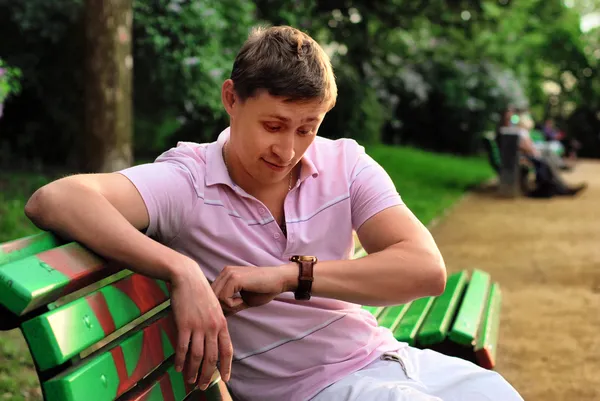 A young man looks at his watch on his arm and sits on a bench in the park and waiting for meeting — Stock Photo, Image