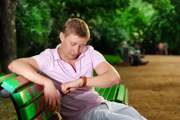 Молодий чоловік дивиться на годинник на руці і сидить на лавці в парку і чекає зустрічі — стокове фото