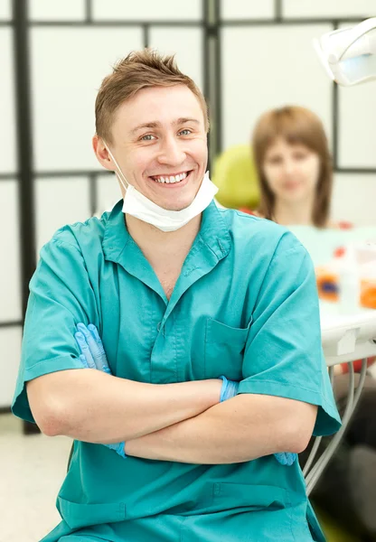 Closeup πορτρέτο του νέους ευτυχής οδοντίατρο στην χειρουργική του, κοιτάζοντας προς τον θεατή, στο βάθος του μια Θηλυκός ασθενής — Φωτογραφία Αρχείου