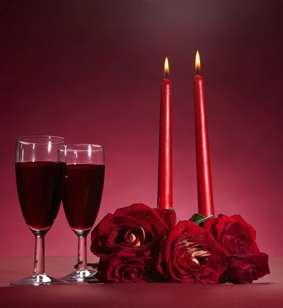 Kytice rudých růží, dvě sklenky vína a dvě svíčky na červeném pozadí — Stock fotografie