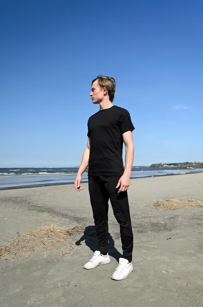 Jeune homme en vêtements noirs et chaussures blanches debout sur la plage — Photo