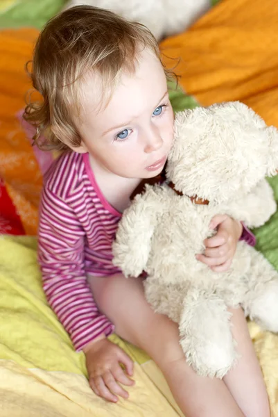 Ledsen liten flicka med blå ögon som sitter på en säng med en nallebjörn. — Stockfoto