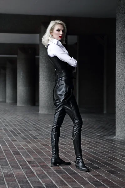 A young blond lady mistress with bright red lips in a white shirt without any emotions wearing a black leather corset and gloves and trousers light behind the model in the streets — Stock Photo, Image