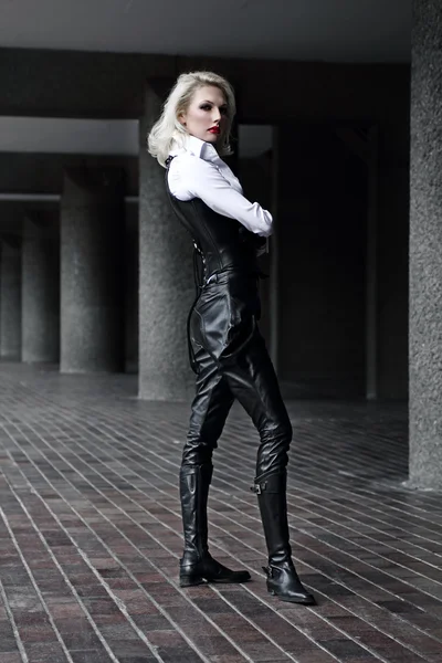 stock image A young blond lady mistress with bright red lips in a white shirt without any emotions wearing a black leather corset and gloves and trousers light behind the model in the streets