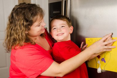Mother and child putting up boy's art on family refrigerator at home clipart