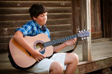 A teenage boy playing a guitar on porch of historic cabin clipart