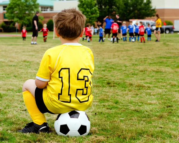 Niño pequeño en uniforme viendo fútbol juvenil organizado o partido de fútbol desde un costado —  Fotos de Stock