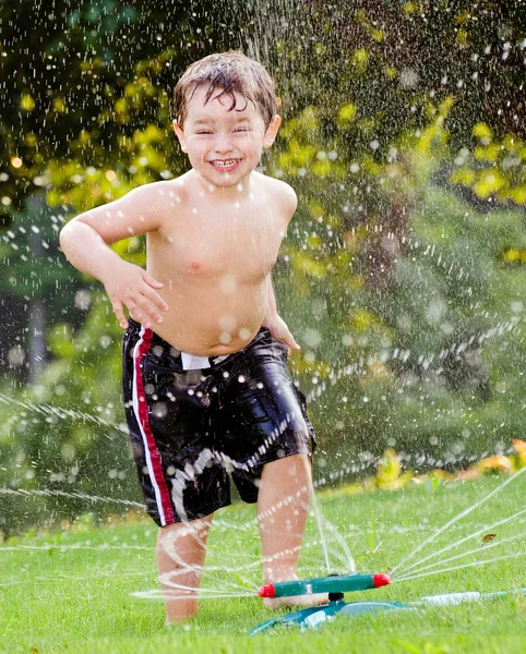 Young boy or kid cools off by playing in water sprinkler at home in his back yard on hot summer day — Stock Photo, Image