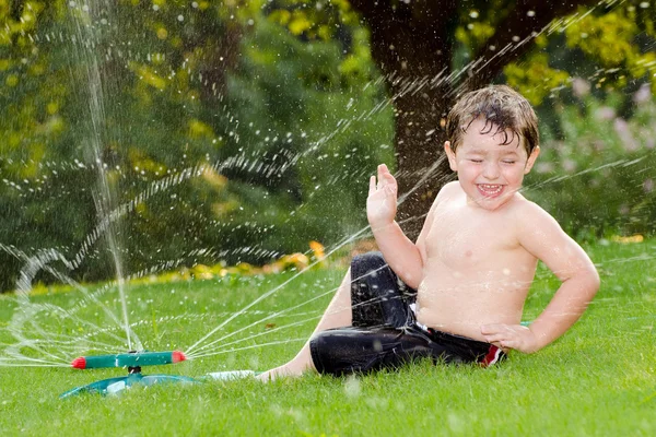 Fiatal fiú vagy gyerek hűti játszva a víz sprinkler otthon a hátsó udvarban, a forró nyári napon — Stock Fotó